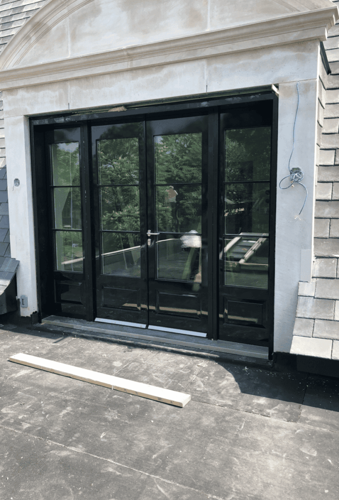 The Armored Embassy Series Double Door features black steel frames, sidelights in a stone wall, visible exterior rooftop plank and wires by the entrance. Trees and greenery are reflected in the glass panels.