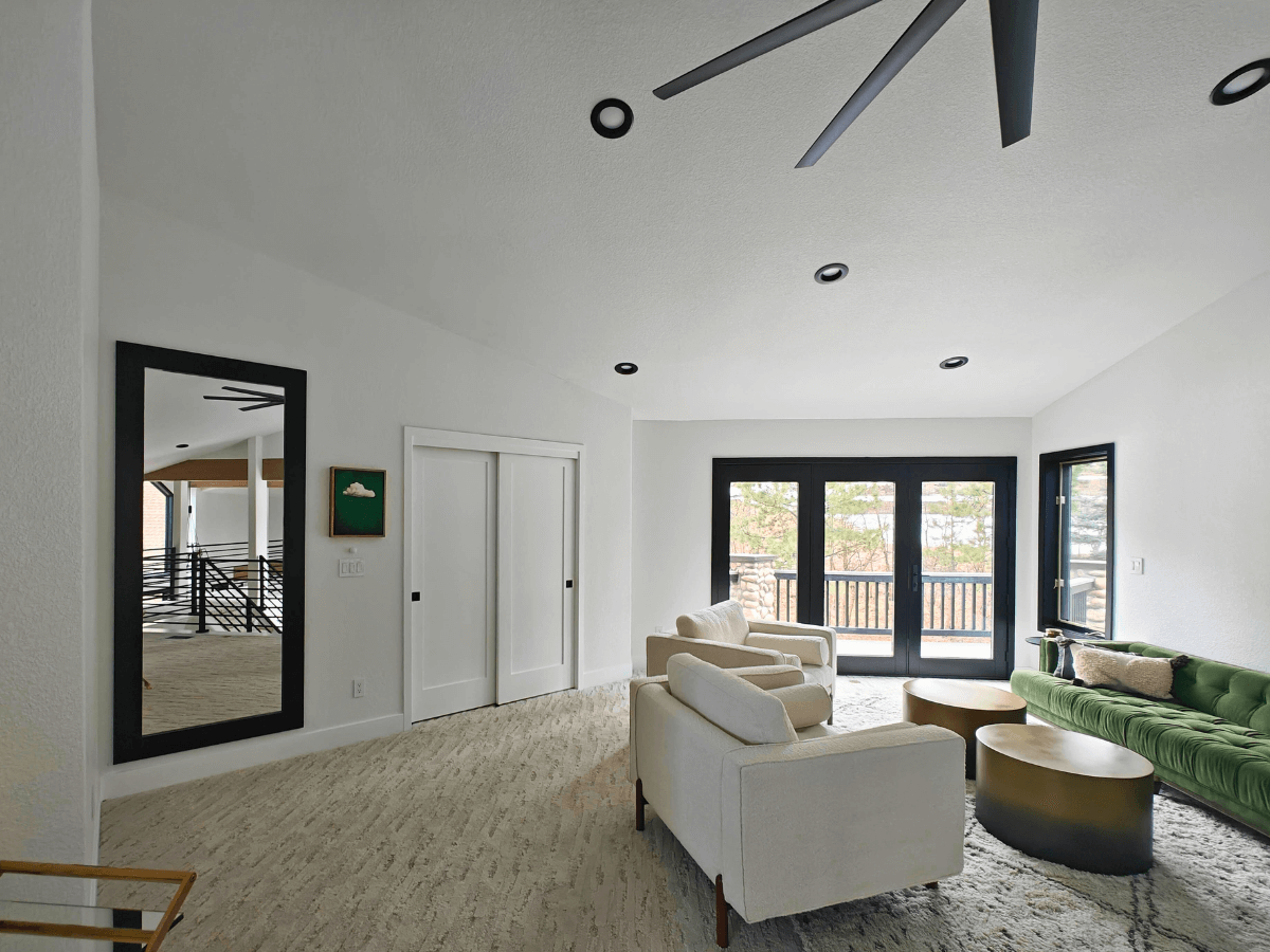 Modern living room with a hidden mirror door on the left wall, white armchairs, a green velvet sofa, and black-framed sliding glass doors leading to a balcony.