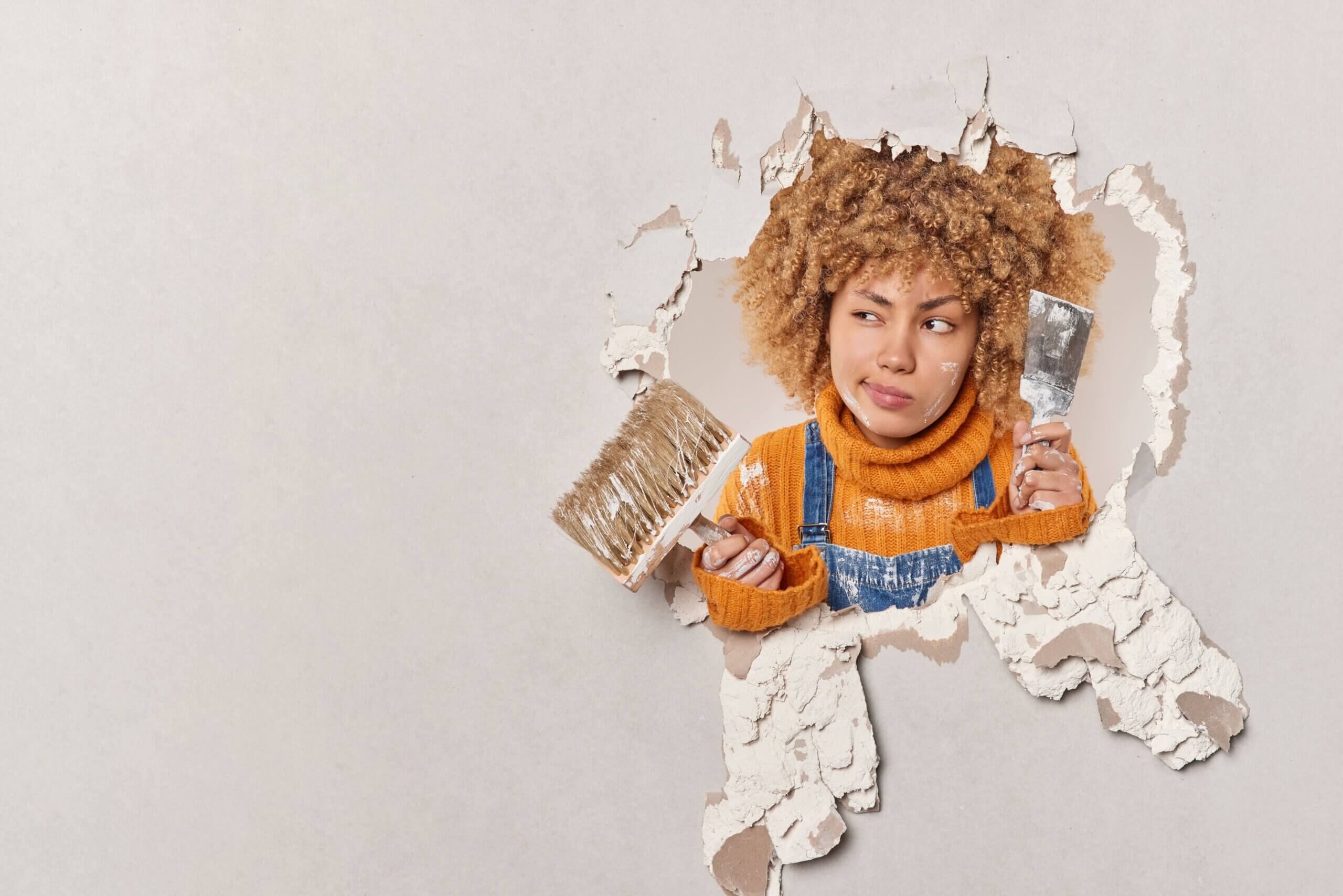 Woman in a sweater and overalls holding a paintbrush and scraper, peeking through a large hole in drywall with a puzzled expression, suggesting a DIY renovation mishap.