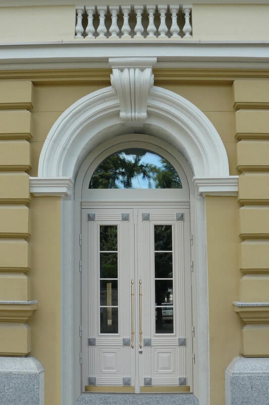  Elegant white double doors with decorative trim and arched transom window on a yellow building facade. Embassy series.