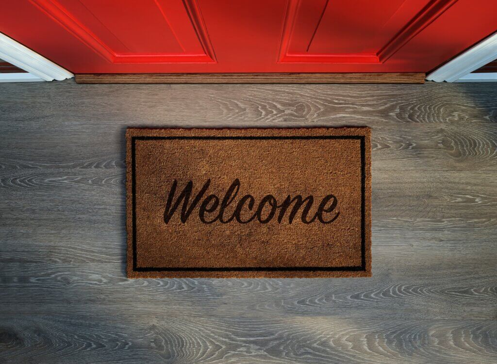 Close-up of a red front door with a brown doormat that says “Welcome” on a wood floor.
