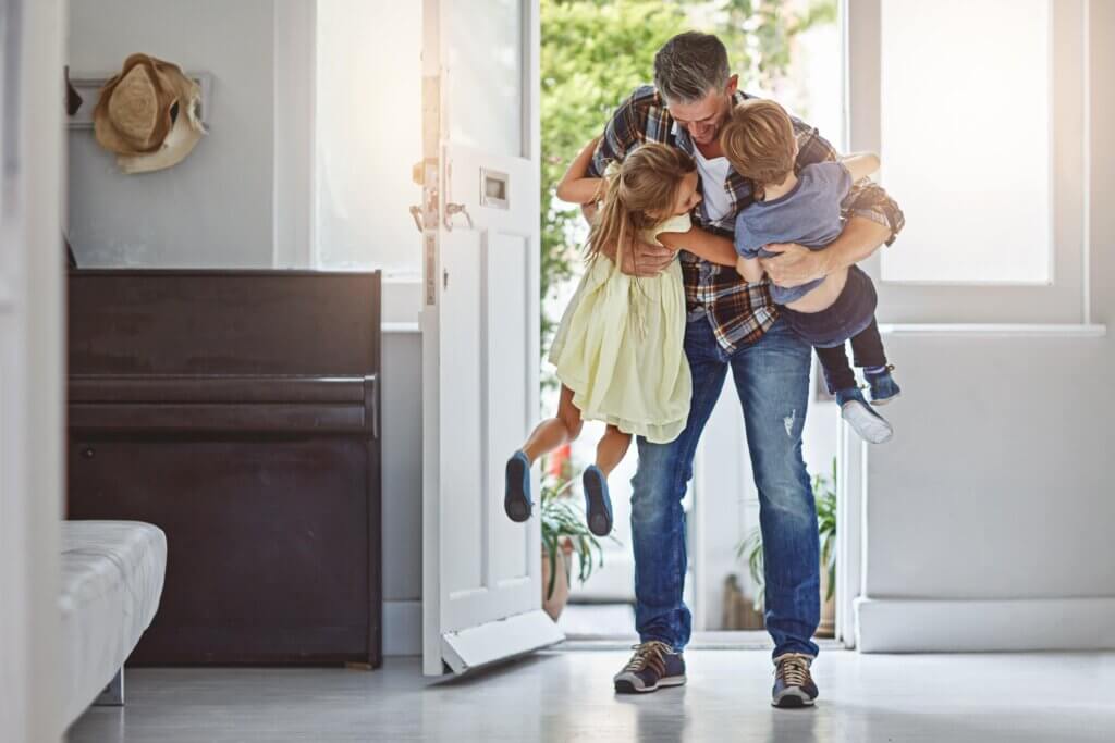 Father warmly hugging two children inside a home entrance with an open white front door.