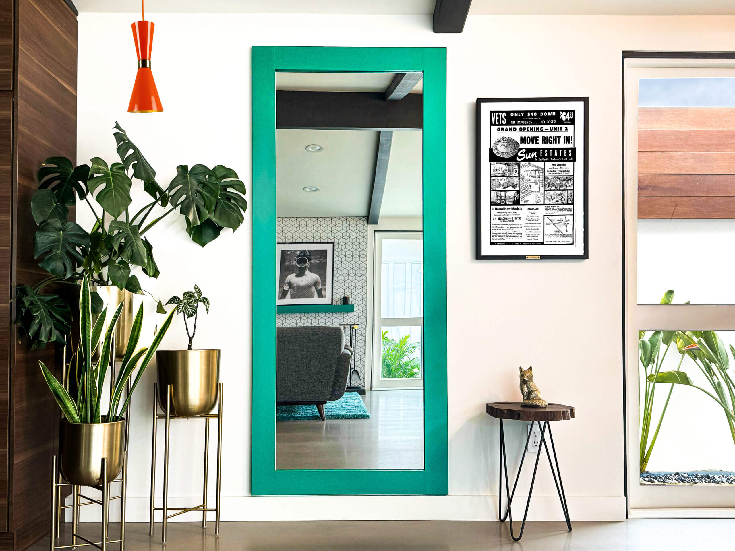 Stylish mid-century modern living room with vibrant teal-framed hidden mirror door, flanked by bold decor and indoor plants. Hidden Door Store.