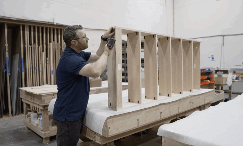 Travis Humble wearing a black polo shirt,  in a workshop, adjusting a hidden bookcase door, with tools and woodworking equipment visible in the background.Testing quality and fit before shipment.