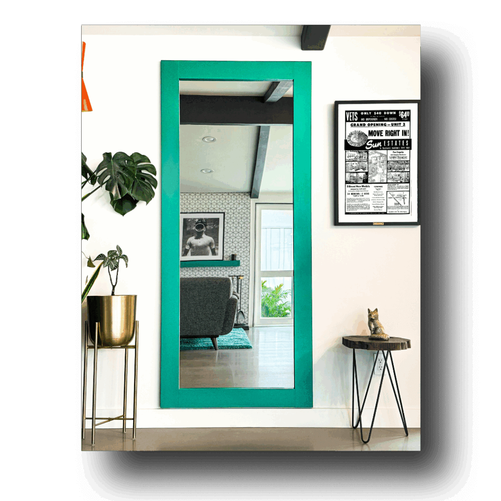  Full-length hidden mirror door with a bold teal frame, reflecting a modern living room with geometric wallpaper, artwork, and natural light from a window, flanked by a potted plant on a gold stand and a small side table with a decorative figure.