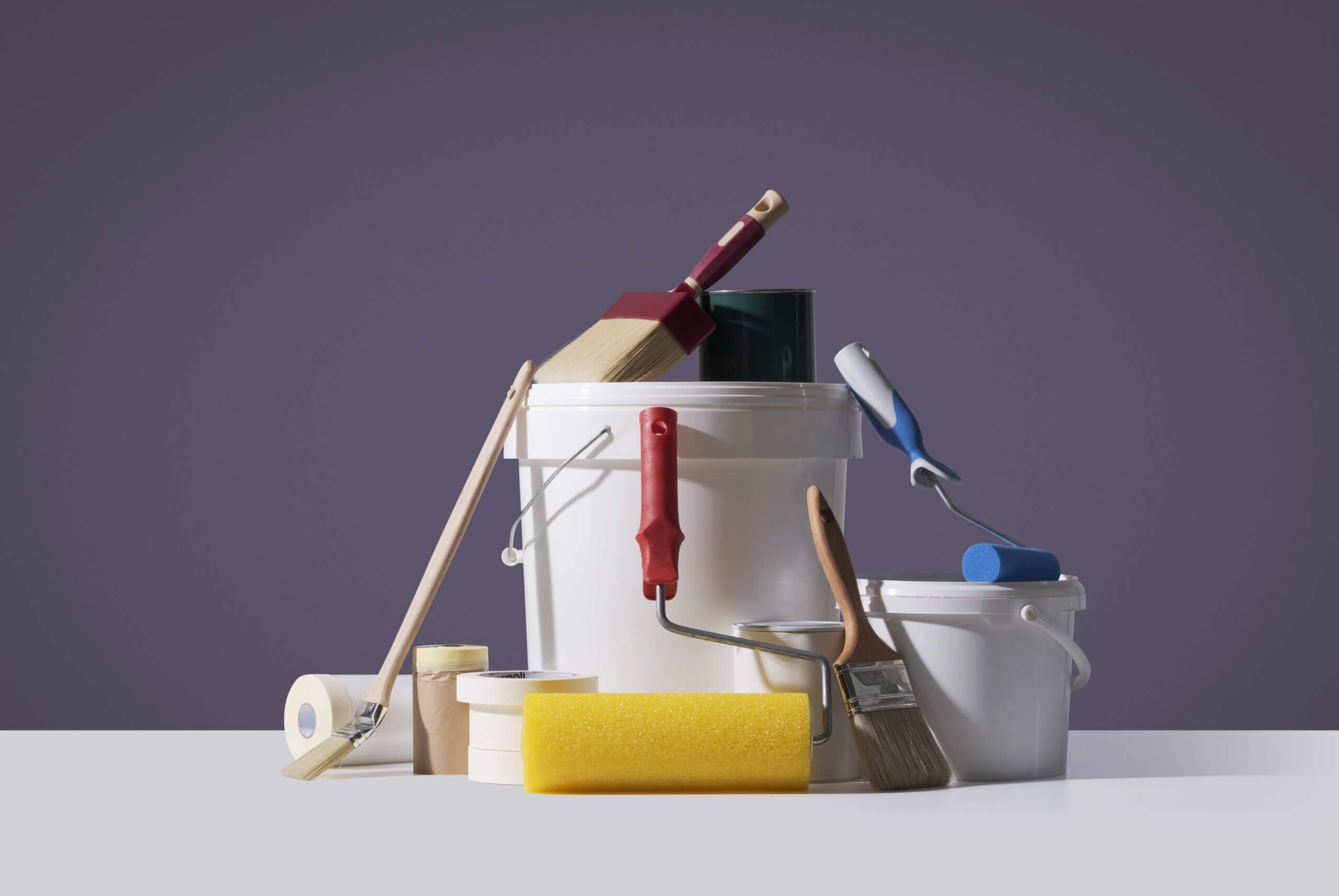 Collection of painting supplies on a white surface with a purple background, including paint buckets, brushes, rollers, masking tape, and a sponge.
