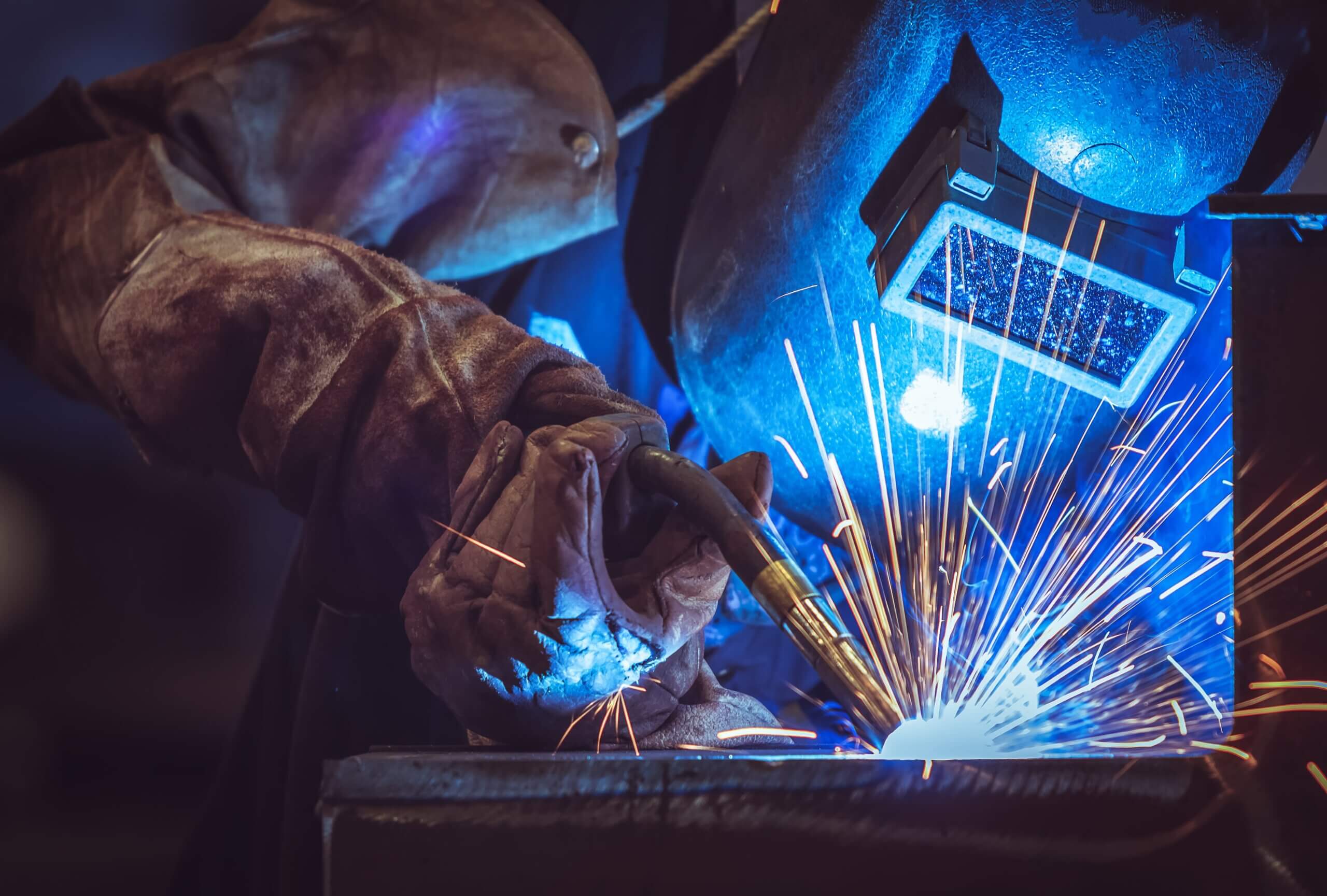 Close-up of a welder in protective gloves and a helmet, creating bright sparks while welding metal, with blue light reflecting off the workspace.