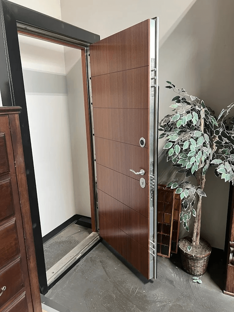 High-security wood-finish steel door manufactured by Hidden Door Store, shown partially open in an interior setting beside a potted tree