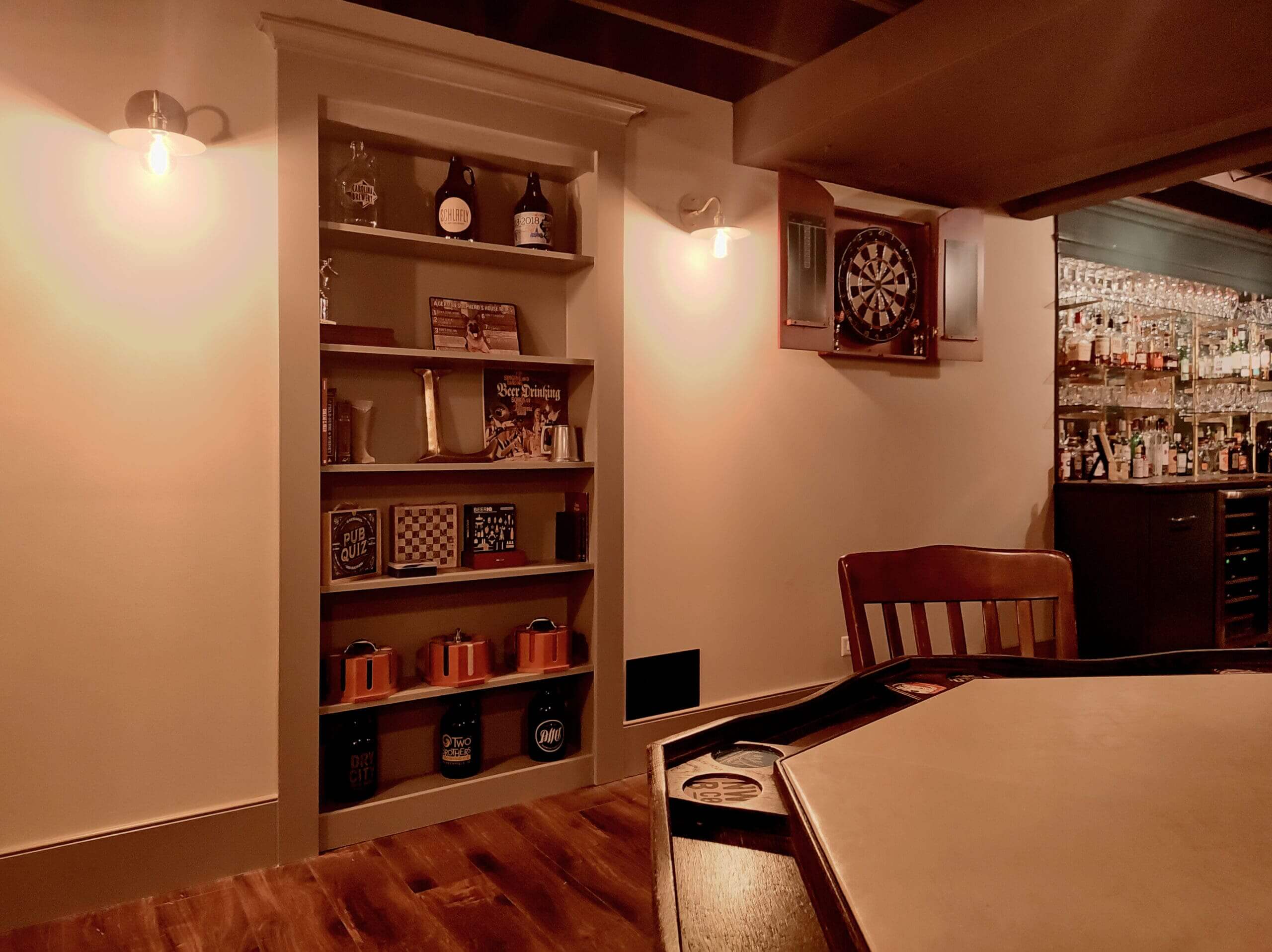 A warmly lit home bar and game room featuring a hidden bookcase door that blends into the wall. The built-in shelves display books, board games, and beer growlers, while a dartboard and mirrored bar stocked with bottles and glassware sit nearby. A wooden chair and gaming table occupy the cozy foreground.