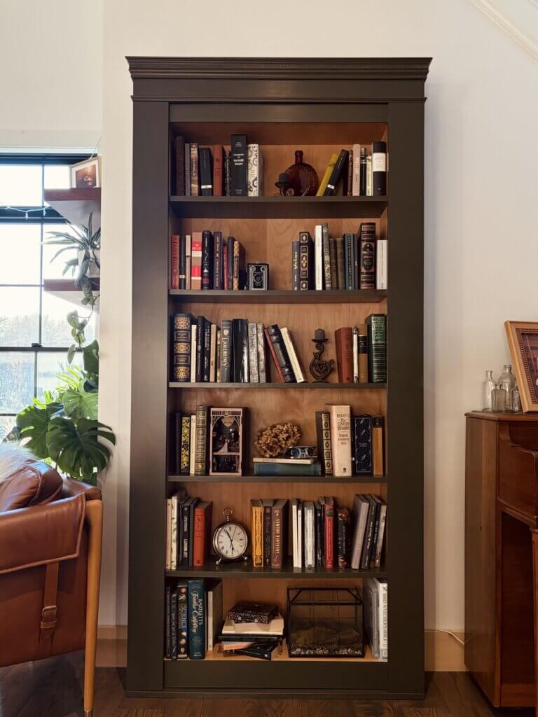 A built-in bookcase filled with books and decorative items, including an antique camera, clock, and glass terrarium. The bookcase has a dark green frame with wood shelving and is positioned between a leather chair and a piano in a cozy, well-lit room.