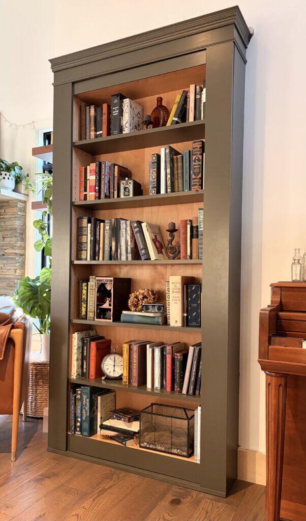  A built-in bookcase filled with books and decorative items, including an antique camera, clock, and glass terrarium. The bookcase has a dark green frame with wood shelving and is positioned between a leather chair and a piano in a cozy, well-lit room.