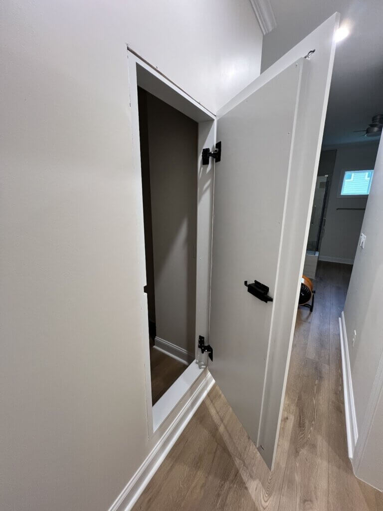 A full-length mirror door seamlessly blends into a hallway wall, reflecting the adjacent closet area with built-in shelving.