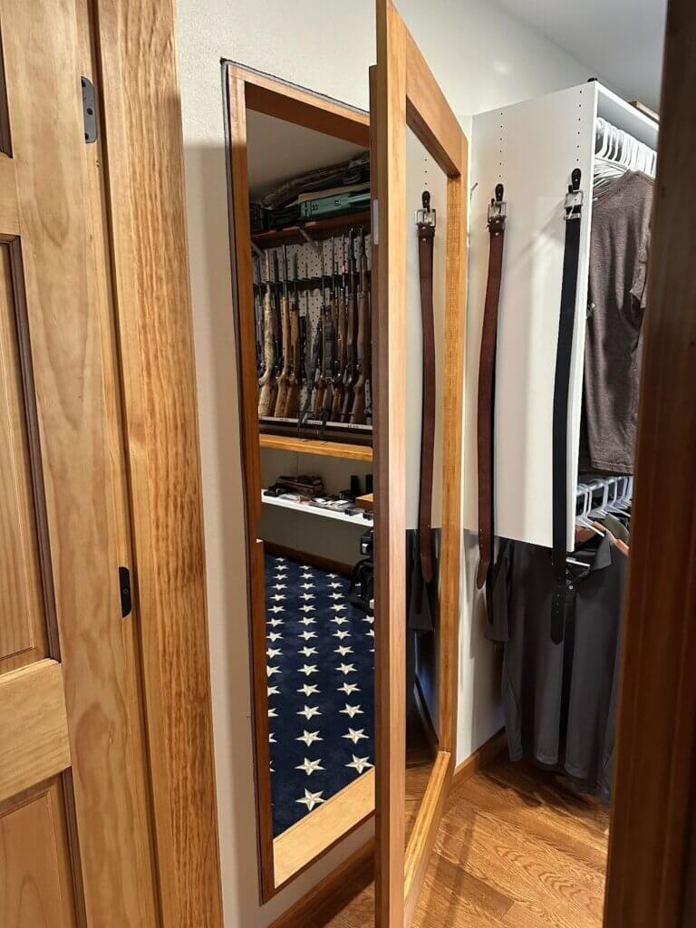A hidden door designed as a full-length mirror opens to reveal a concealed gun room behind it, with rifles stored on a pegboard wall and a blue carpet with white stars on the floor.