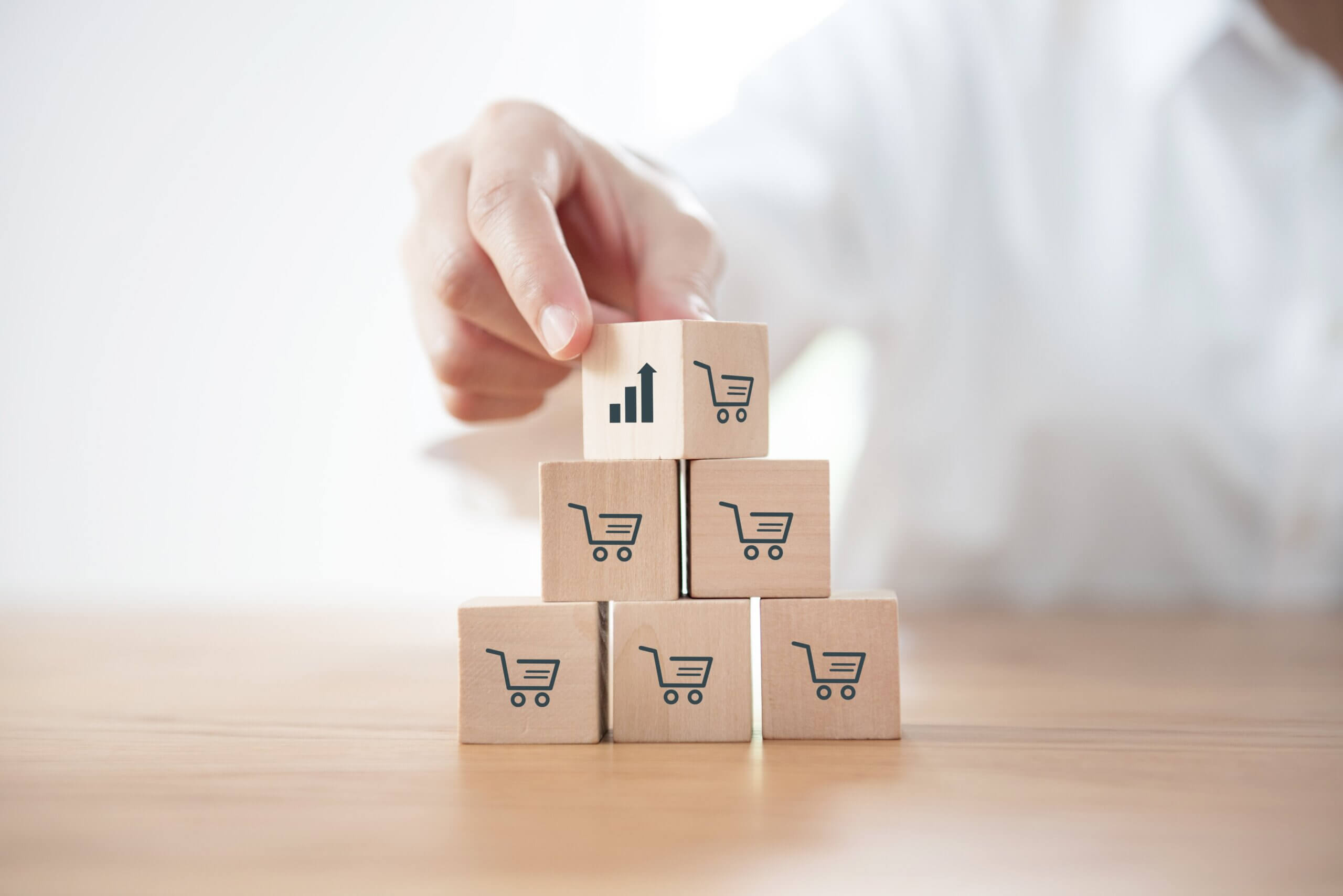 A person stacking wooden blocks with shopping cart icons, symbolizing e-commerce growth. The top block features an upward arrow and bar chart icon, representing direct to consumer sales.