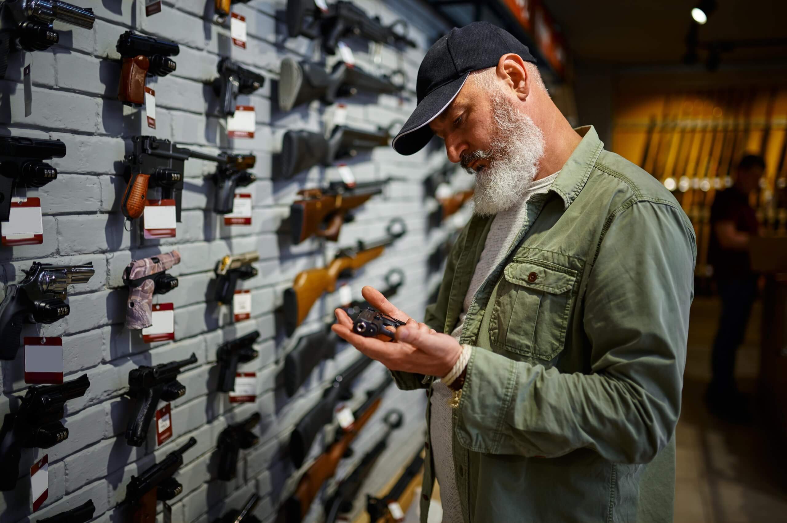 An older man with a white beard and a black cap examines a small handgun in a gun store, standing in front of a display wall lined with various pistols and rifles.