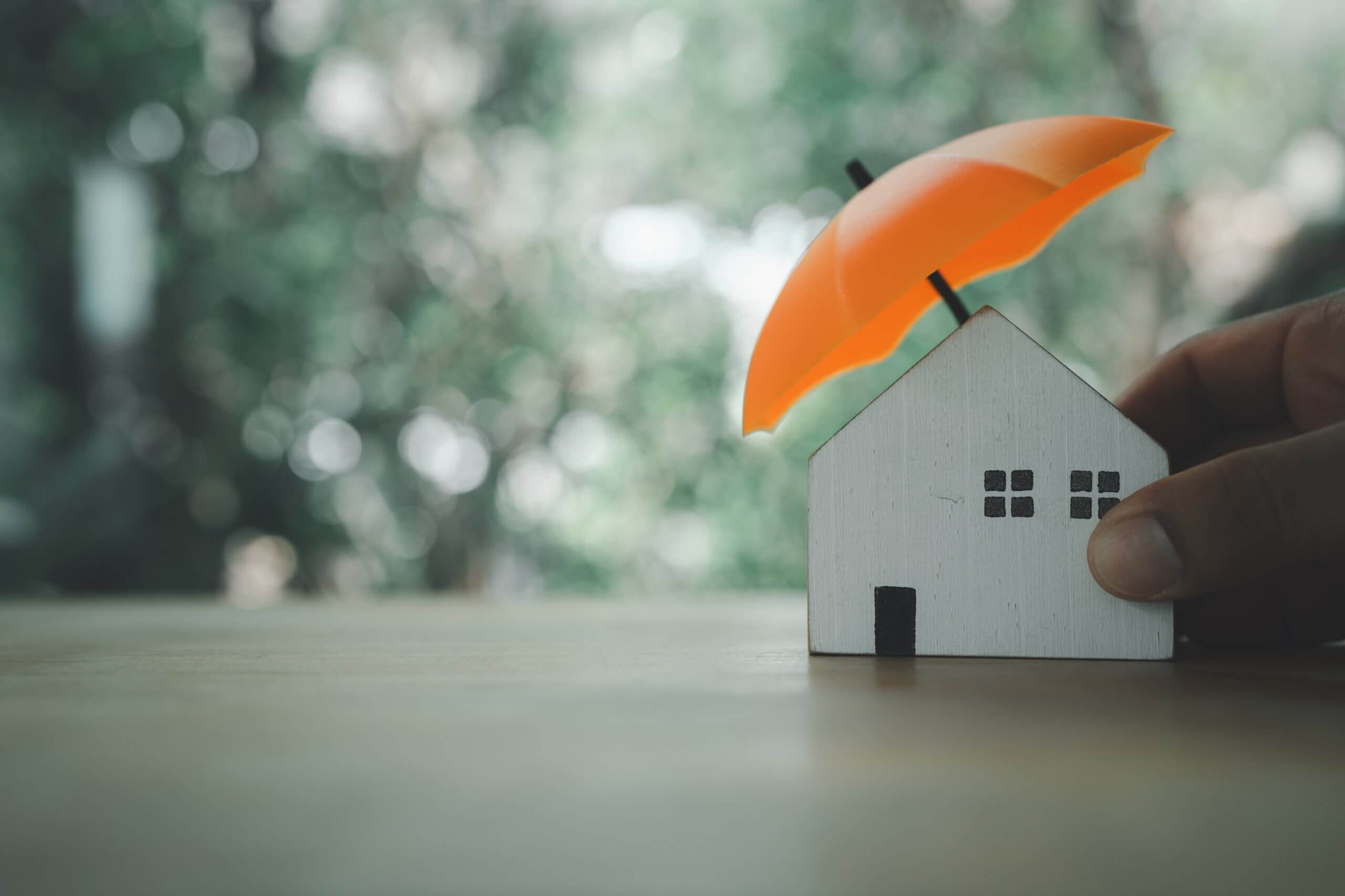Hand holding a small house model with an orange umbrella symbolizing home protection and security