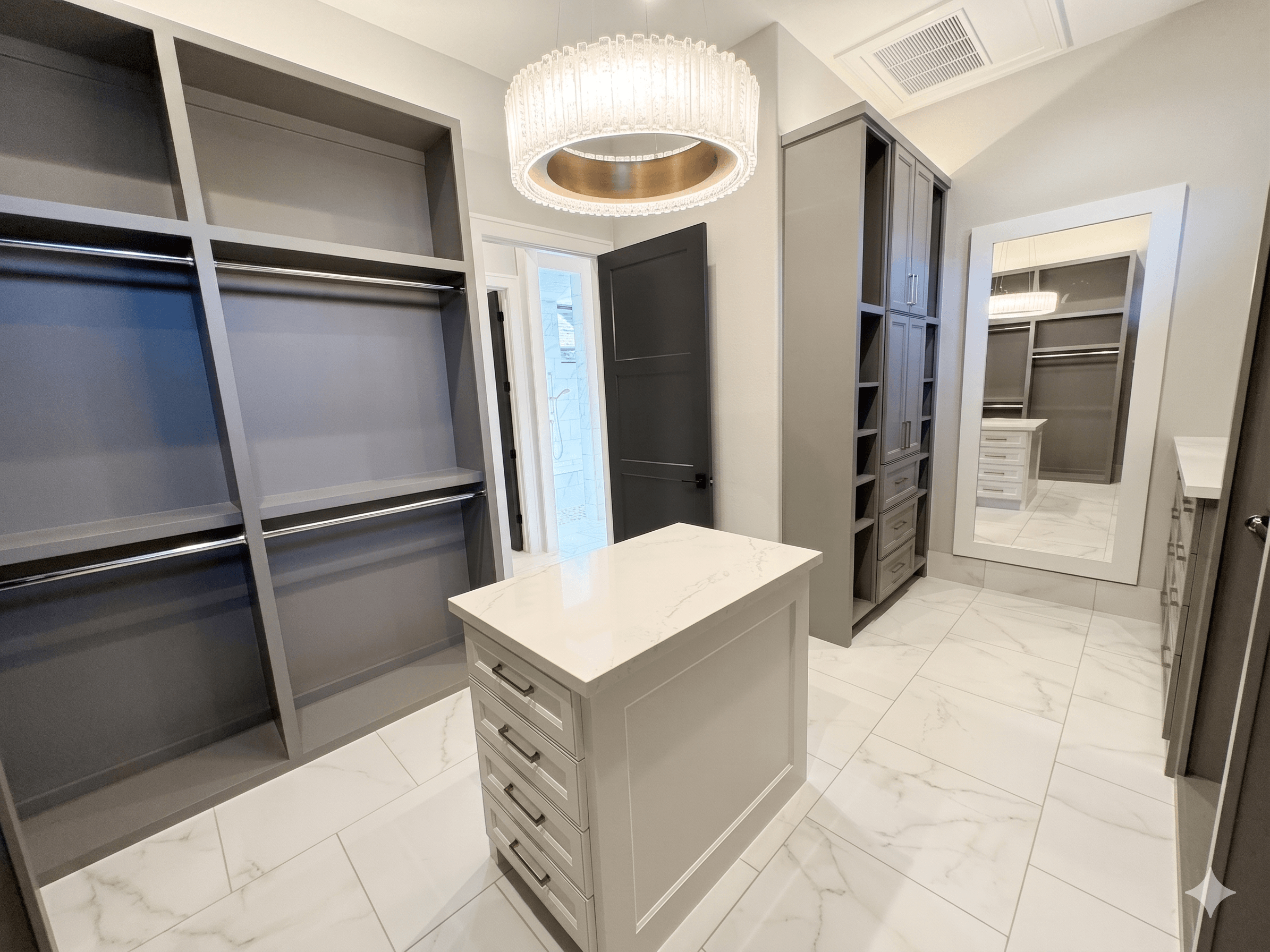 Spacious luxury walk-in closet with gray custom built-in shelving, marble tile flooring, center storage island with drawers, large full-length hidden mirror door, and modern crystal chandelier lighting.