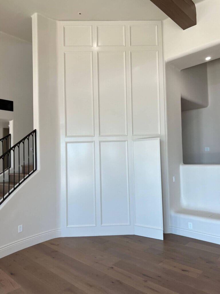 Modern white hidden door seamlessly blended into tall paneled wall design in a luxury home with wood flooring and curved architectural details. 